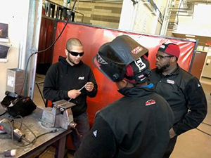 man shows welding piece to two welding students