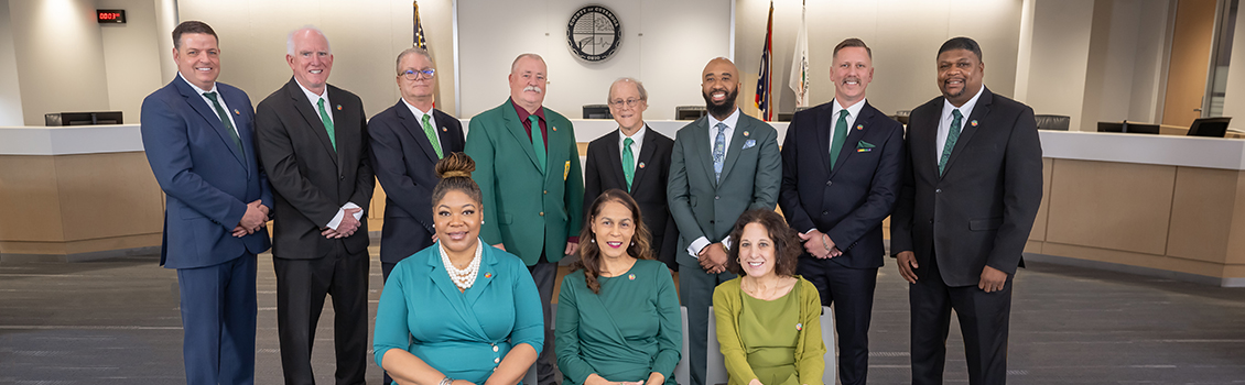 Council Group of eleven professionally dressed people posing together in a conference or council room with a large clock and flags in the background.