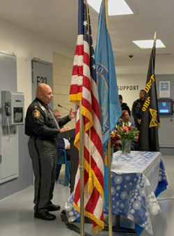 Man in sheriff's uniform standing at a podium with U.S. flags in the foreground.