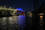 A bridge span over water at night is illuminated in red, white, and blue lighting