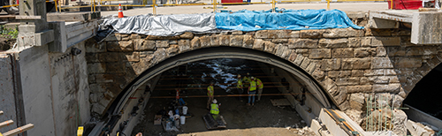 A stone bridge with tarps, construction workers in safety vests, and some yellow security barriers.