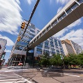 Amodernsilverandglassofficebuildingwithasuspendedwalkwayoveracityintersectionwithtrafficlightsandcarspassingthrough