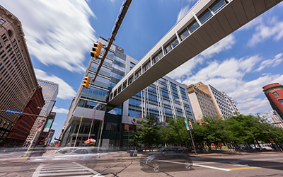 Amodernsilverandglassofficebuildingwithasuspendedwalkwayoveracityintersectionwithtrafficlightsandcarspassingthrough