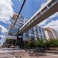 Amodernsilverandglassofficebuildingwithasuspendedwalkwayoveracityintersectionwithtrafficlightsandcarspassingthrough