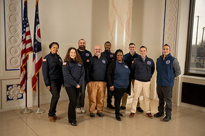 A group of professionals is standing together smiling, each wearing a pullover with an Emergency Management Agency patch.