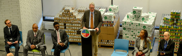 A man in a suit stands at a podium with a county logo on it in front of people in chairs.