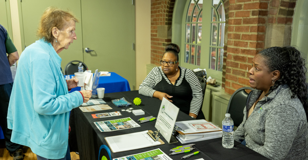 An older adult is standing in front of a table talking to two women who are seated behind the table. There are many brochures and pamphlets on the tabletop.