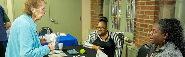 An older adult is standing in front of a table talking to two women who are seated behind the table. There are many brochures and pamphlets on the tabletop.