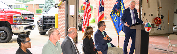 Crisis Response Program Expands A man in a business suit is standing at a podium and speaking while a group sits in chairs nearby and listens.