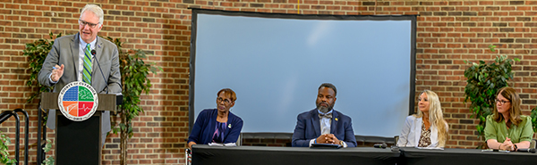 CSU A man in a business suit is standing at a podium and speaking, while four people listen and are seated in a table to his left.