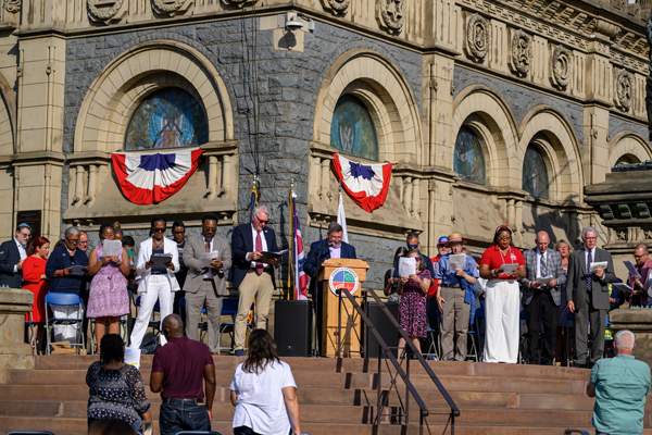 A man standing at a podium reading out loud in front of a large building surrounded by people.