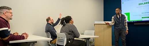A man stands before a screen and by a podium before three adults sitting at tables and one is raising his hand.
