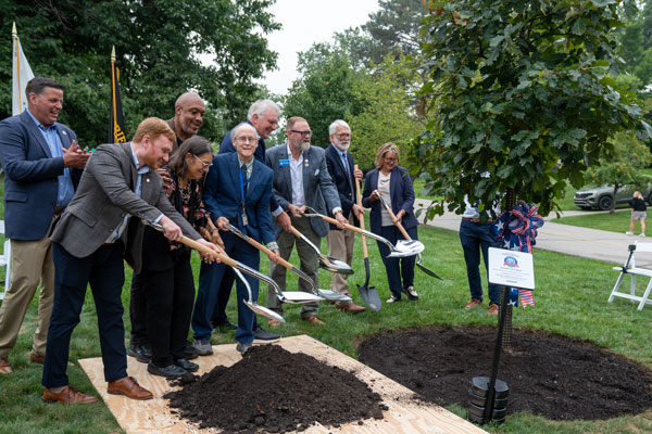 A group of adults in business clothes shoveling dirt onto a newly planted tree in the ground.
