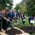 Agroupofadultsinbusinessclothesshovelingdirtontoanewlyplantedtreeintheground