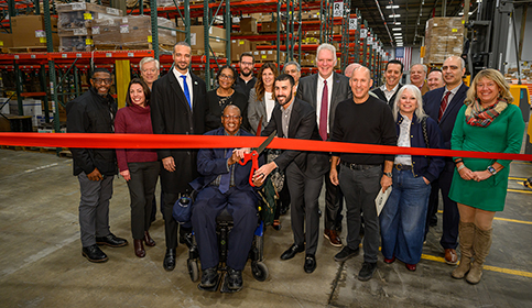 BusinessLending-FamousSupply Group of people in warehouse during a ribbon cutting ceremony cutting ribbon with large scissors.