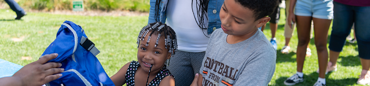 kid getting a backpack at Family Fun Day – A person is handing a new blue backpack to a child who is smiling with another older child looking on