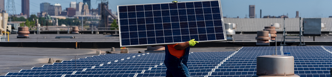 A worker is carrying a solar panel and walking on a roof with a solar panel array