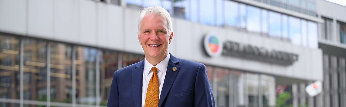 Chris Ronayne and Cuyahoga County headquarters Smiling man in a blue suit and orange tie standing outside a modern building with glass windows and the Cuyahoga County logo in the background.