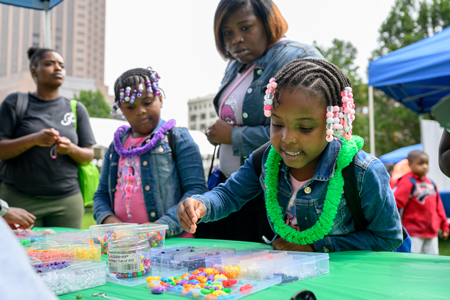 A young child wearing a green plastic lei is selecting colorful beads from a table with another child and adult.