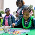 Ayoungchildwearingagreenplasticleiisselectingcolorfulbeadsfromatablewithanotherchildandadult