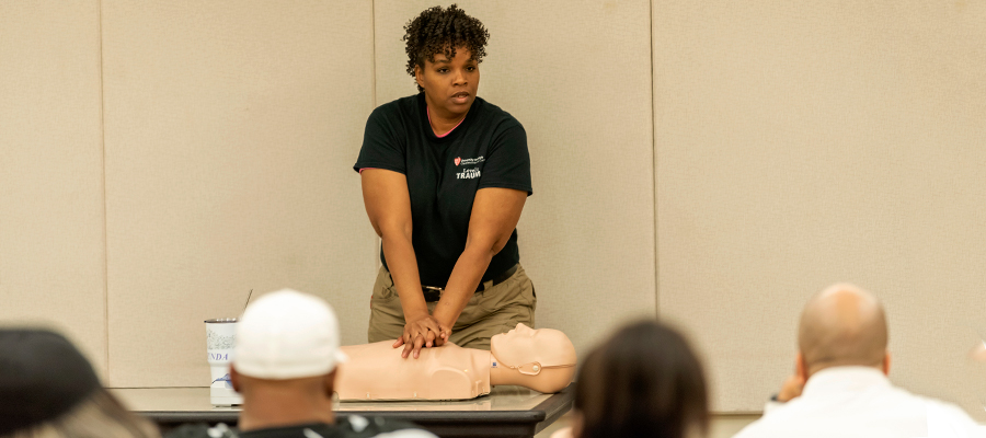 A woman giving chest compressions to a dummy in front of a class.