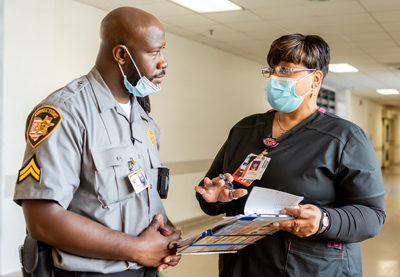 A Corrections Officer speaking with a woman wearing a surgical mask and hospital scrubs.