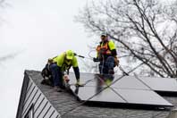 Two workers wearing safety gear are on top of a house are attaching solar panels.