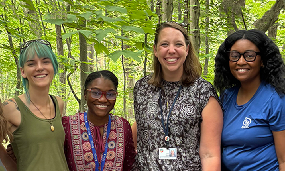 Four young women standing and smiling together in the woods.