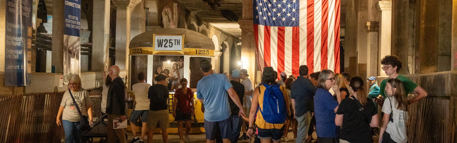 A crowd of people looking at a historic yellow subway with an American flag hanging from the ceiling.